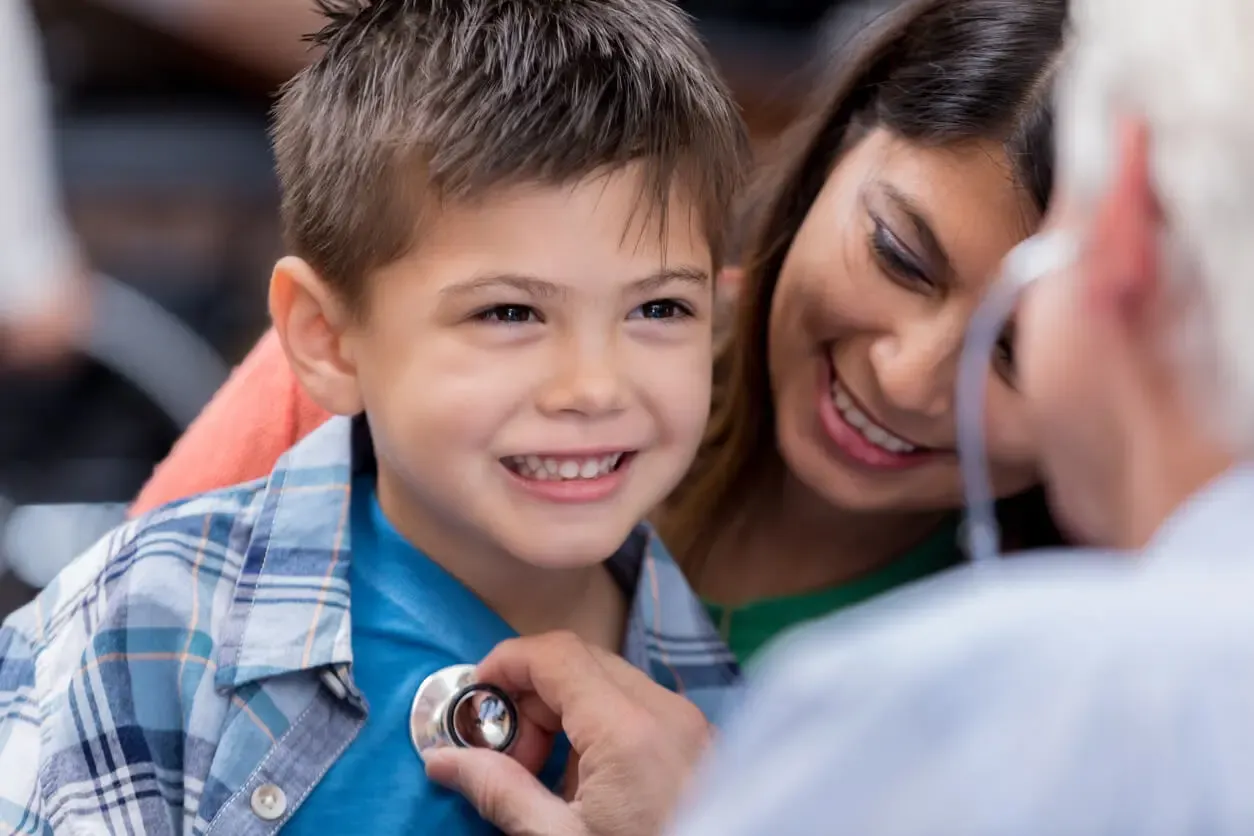 A smiling female healthcare provider kneeling to talk with a young child patient, holding a stethoscope, in a bright pediatric exam room.