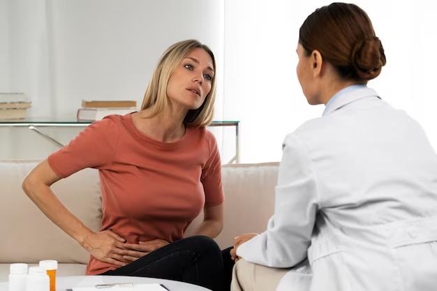 A woman in a red shirt sits on a couch, holding her stomach, while speaking to a female doctor in a white coat at the medical center.