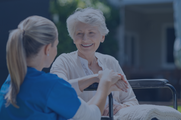 A smiling elderly woman in a wheelchair is outdoors, engaging in conversation with a healthcare worker in a blue uniform from the nearby urgent care.