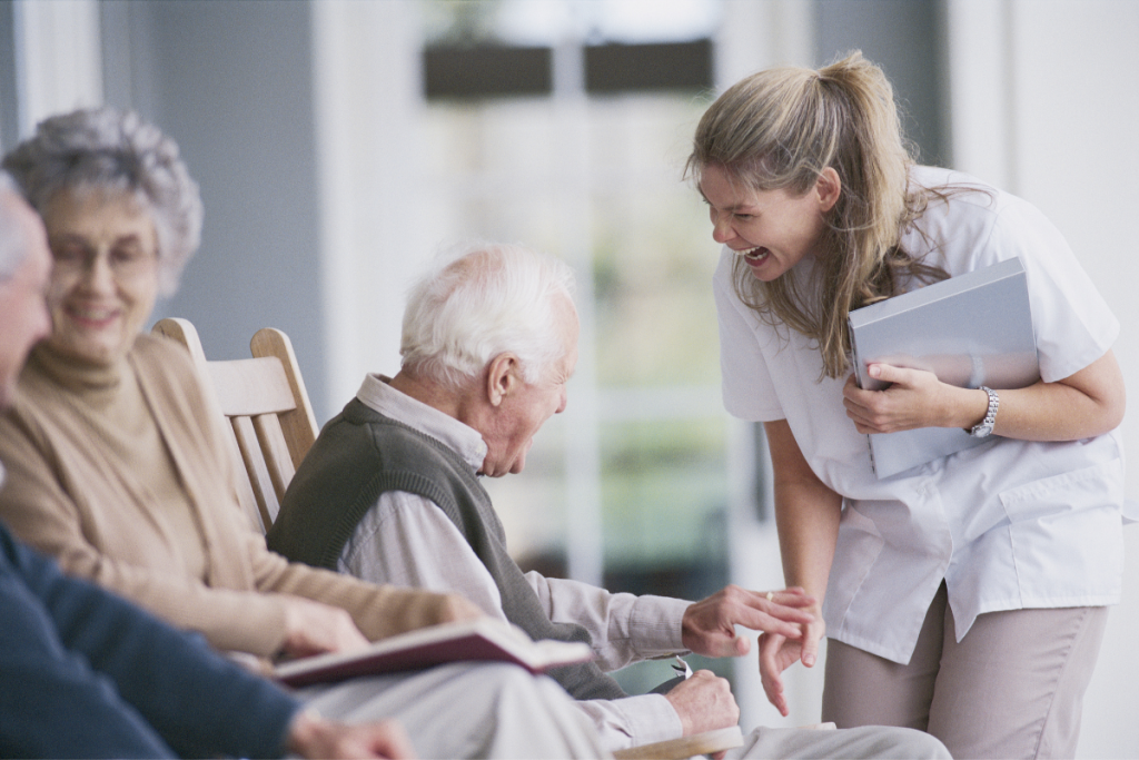 A group of elderly people sit on chairs, with one elderly man smiling and interacting with a female caregiver who is holding a folder and laughing at the medical center.