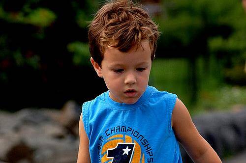 A young boy with short, light brown hair, wearing a blue sleeveless shirt, looks down with a serious expression. Behind him, the background is blurred with greenery, resembling the surroundings of an urgent care clinic.
