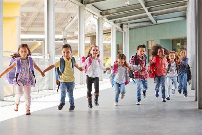 A group of young children with backpacks run and hold hands together down an outdoor walkway near the local walk-in clinic.