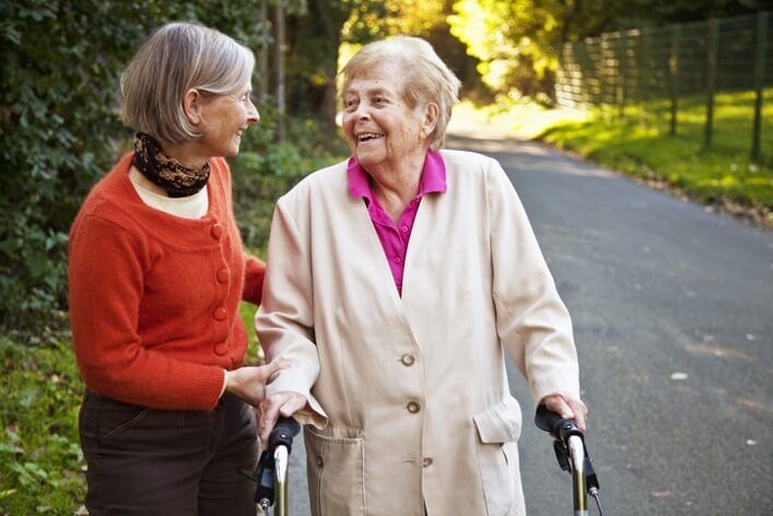 Two elderly women walking outside on a pathway; one woman uses a walker and the other woman is supporting her. They are smiling and surrounded by green foliage, having just enjoyed a visit to the walk-in clinic nearby.