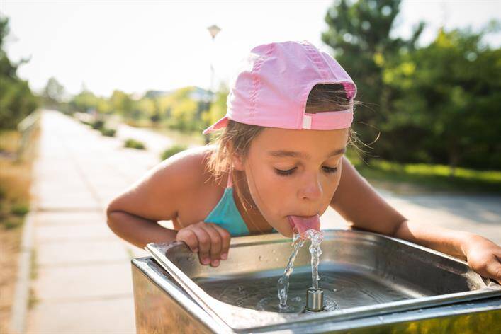 A young girl wearing a pink cap drinks water from a public drinking fountain on a sunny day, right next to the local medical center.