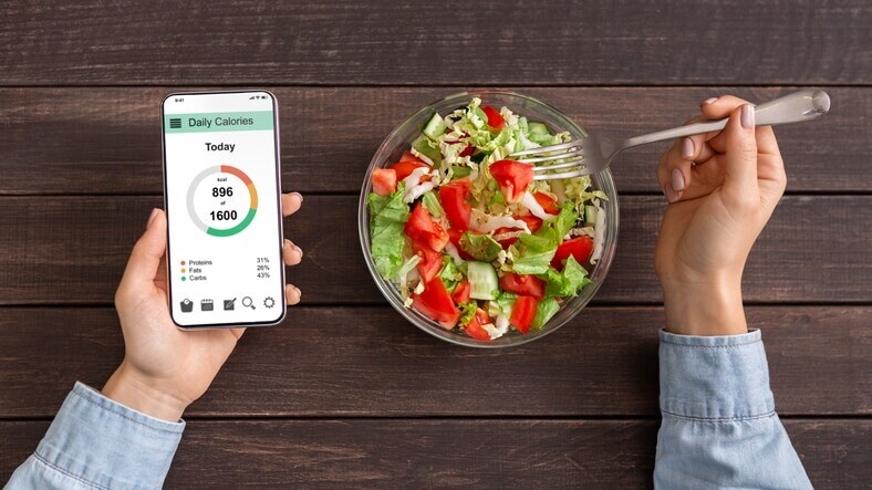 A person, fork in hand, eats a bowl of salad with lettuce, tomatoes, and other vegetables on a wooden table while using a smartphone displaying a calorie tracking app—reminiscent of tools found in medical centers or urgent care facilities.