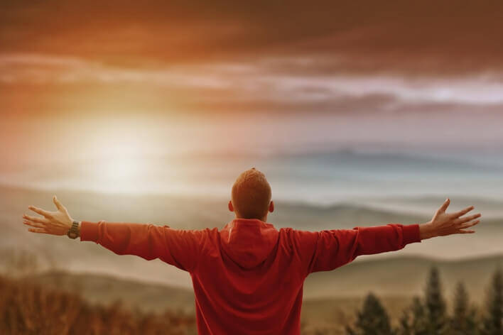 Person in a red hoodie standing with open arms, facing the sunrise over a mountainous landscape, finds solace after a late-night visit to the urgent care.