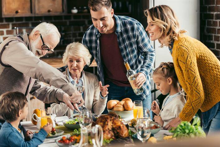 A family of six, spanning three generations, gathers around a dining table set with a variety of dishes, including a roast turkey, while two adults share stories about their recent visit to an urgent care clinic.