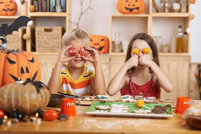 Two children holding Halloween-themed decorations in front of their eyes, sitting at a table with pumpkins, cookies, and festive decor, could easily brighten up a visit to any walk-in clinic this spooky season.
