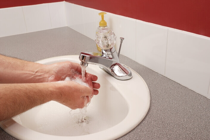 A person washing their hands with soap under running tap water in a bathroom sink at an urgent care facility. A bottle of liquid soap is placed beside the tap on a grey countertop.