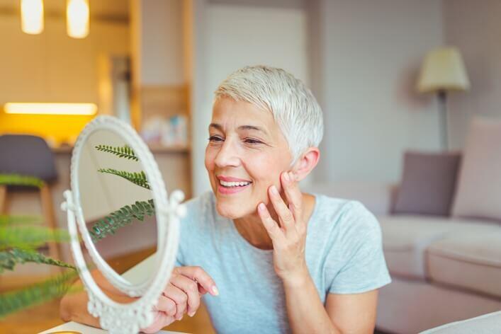 An elderly woman with short gray hair smiles as she touches her cheek and looks into an ornate hand mirror in a cozy, well-lit living room, recalling her recent visit to the walk-in clinic.