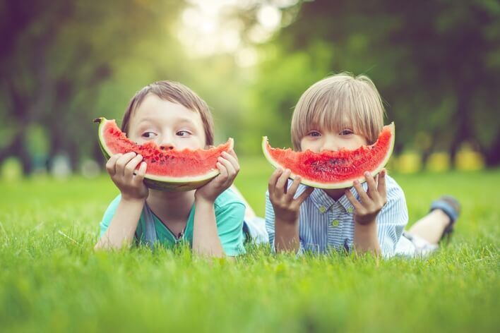 Two children lie on the grass, each holding a slice of watermelon close to their faces. The background is a blurred green park setting with trees, making it seem like a perfect spot you'd visit after dropping by an urgent care walk-in clinic for a quick check-up.