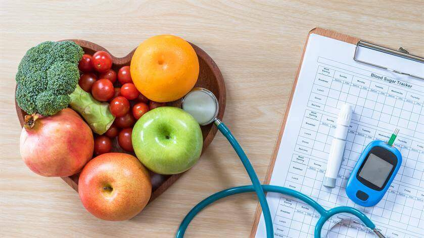 A heart-shaped bowl filled with fruits and vegetables, alongside a stethoscope, blood sugar tracker, and clipboard on a wooden surface, captures the holistic approach of an urgent care setting.