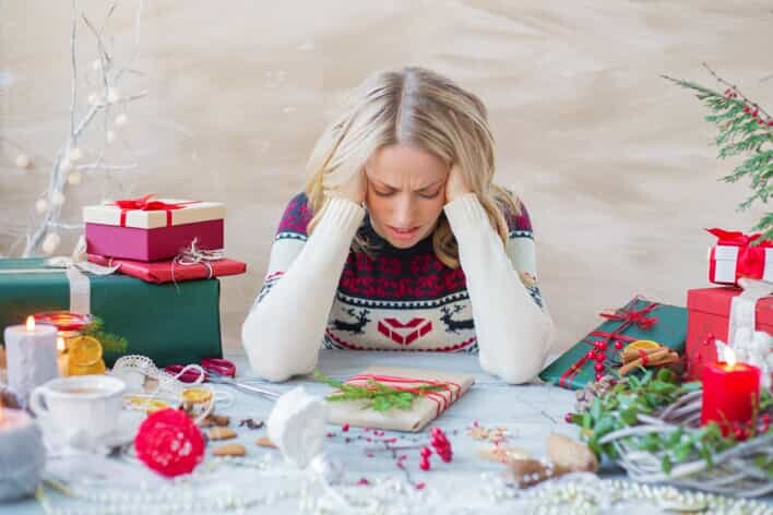 A woman in a holiday-themed sweater looks down with a frown, surrounded by wrapped Christmas gifts, candles, and festive decorations on a table, as though she just received an urgent call from the medical center.