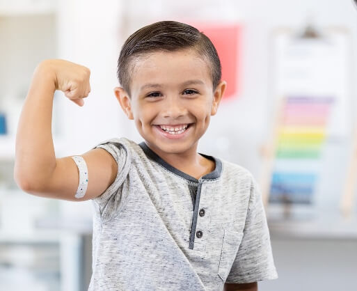 A young boy with a bandage on his upper arm flexes his bicep and smiles, indicating he has received a vaccination at the medical walk-in clinic.