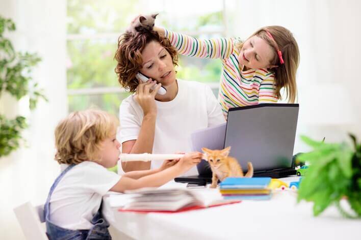 A woman is on the phone with two children around her; one child is brushing her hair with a toy, and the other plays with a kitten on the table near a computer, while she arranges an appointment at the urgent care center.