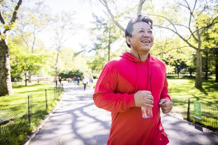 A person in a red hoodie is jogging in a park, holding a water bottle and wearing earphones, with trees and sunshine in the background, seemingly heading toward the nearby urgent care clinic.