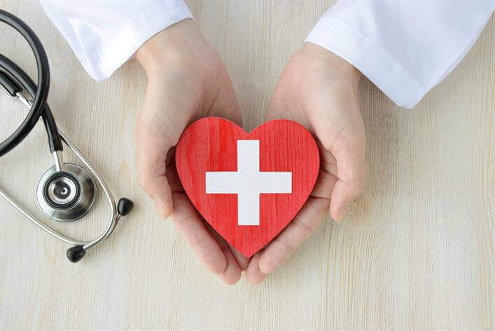 A person in a white lab coat holding a red heart-shaped object with a white cross in the center. A stethoscope lies on the table nearby at the walk-in clinic.