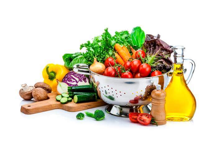 A variety of fresh vegetables, including tomatoes, lettuce, carrots, and mushrooms, arranged around a colander with a bottle of olive oil and a pepper grinder on a white background—ideal for promoting healthy eating in any medical center.