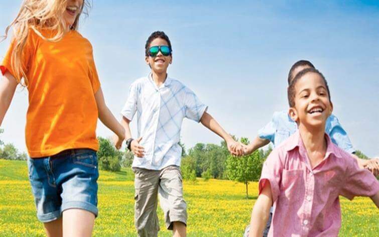 Four children are running hand-in-hand through a field of grass and yellow flowers under a blue sky, the scene so carefree you’d never know that just hours before, they were at an urgent care clinic for minor scrapes.