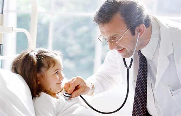 A doctor at the medical center uses a stethoscope to check the heartbeat of a smiling young girl lying in a hospital bed.