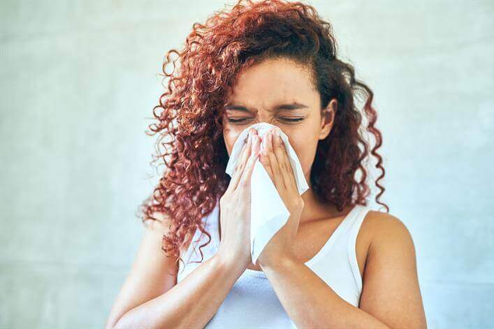 A person with curly hair blowing their nose into a tissue while closing their eyes. They are wearing a white sleeveless top, standing against a light-colored background that resembles the clean atmosphere of an urgent care center.