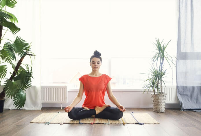 A person is meditating in a cross-legged position on a mat in a bright room with large windows and several green plants, finding peace amidst the hustle of daily life, much like the serene moments sought after visiting an urgent care walk-in clinic.