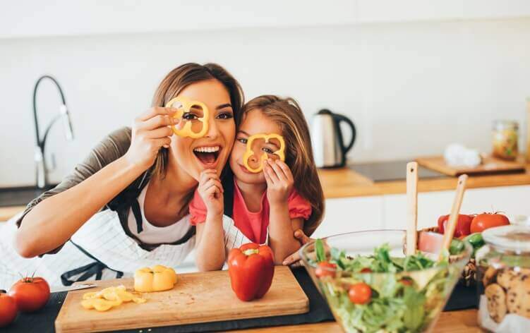 A woman and a young girl are in a kitchen, holding slices of bell pepper in front of their eyes and smiling. A salad bowl and other vegetables are on the counter, creating an atmosphere much more inviting than any visit to a medical center or walk-in clinic.