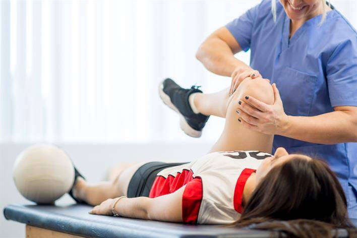 A person in sportswear lies on a table while a medical professional in blue scrubs performs a physical therapy session, focusing on the person's knee. Nearby, at the walk-in clinic's facility, a volleyball rests beside the individual.