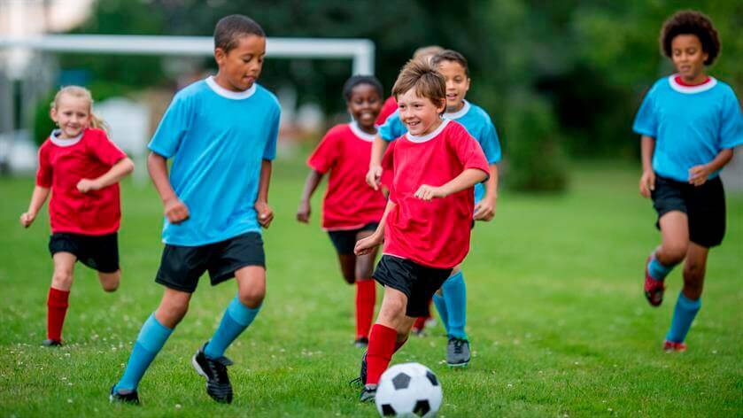 Children wearing red and blue soccer kits are running and playing a game of soccer on a grassy field, with an urgent care clinic nearby for any unexpected medical needs.