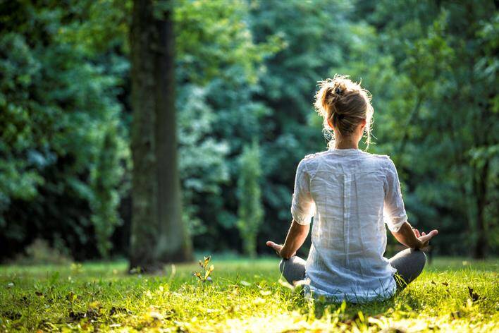 Person sitting cross-legged on grass in a park, near the medical center, facing away from the camera, meditating with hands resting on knees.