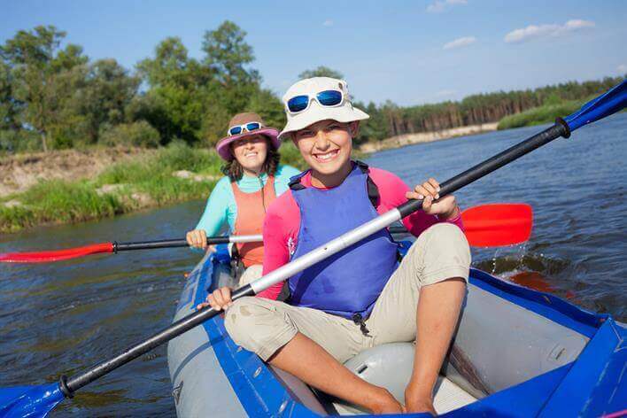 Two people are canoeing on a calm river, both wearing hats, sunglasses, life jackets, and holding paddles. Trees and a clear sky provide the backdrop, creating a tranquil escape perfect for unwinding after a long week at the medical center.