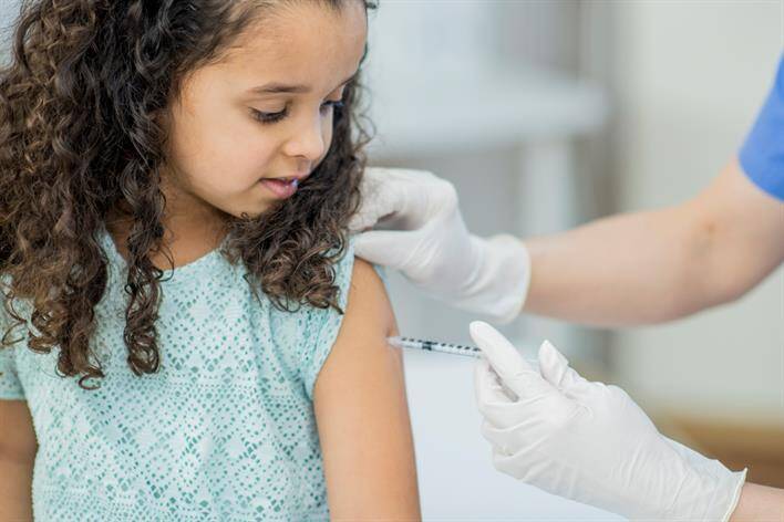 A healthcare professional at a walk-in clinic gives a young girl an injection in her upper arm while the girl looks away.