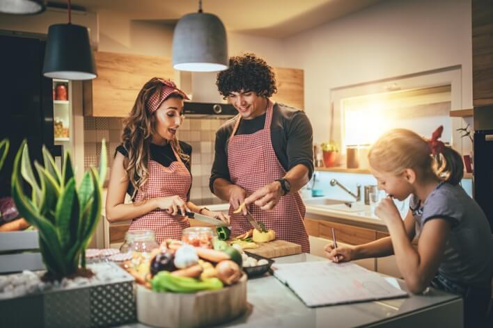A man and woman, both in aprons, are chopping vegetables in a modern kitchen, while a young girl sits at the counter, watching and writing in a notebook. The scene could be mistaken for an urgent care simulation rather than their peaceful family evening.