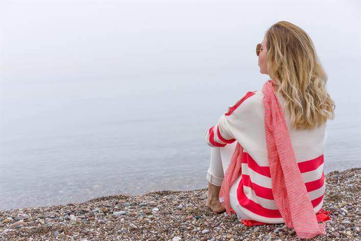 A person with long hair, wearing a red and white striped sweater, sits on a rocky beach facing a foggy, calm sea. Nearby, the faint outline of an urgent care center emerges through the mist.