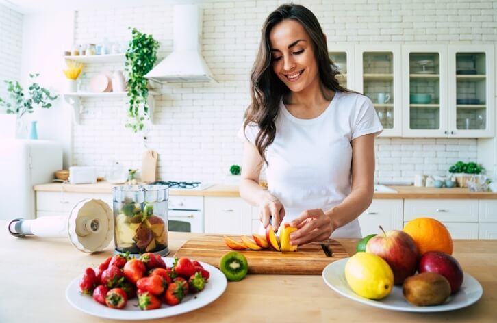 A woman in a white t-shirt is smiling as she slices fruit in a bright kitchen, with plates of various fruits and a blender on the counter, resembling the warm welcome you'd get at an urgent care center.