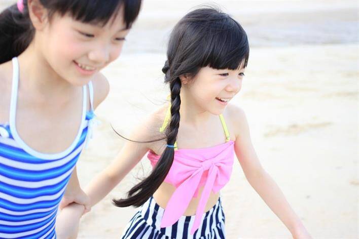 Two children, one in a blue striped swimsuit and the other in a pink top with striped bottoms, holding hands and smiling while walking on a sandy beach near the medical center.