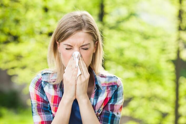 A woman in a plaid shirt is sneezing into a tissue outdoors in a green, leafy setting, possibly on her way to a walk-in clinic.