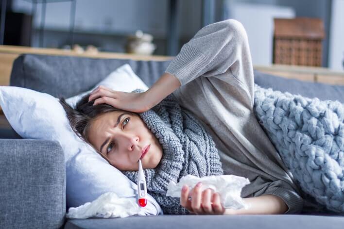 A person lying on a couch with a thermometer in their mouth, holding tissues, and appearing unwell, considering a visit to the urgent care.