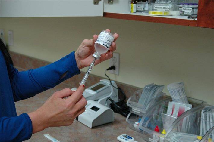 A person is drawing a liquid from a vial into a syringe in a walk-in clinic, with medical supplies organized on the countertop in the background.