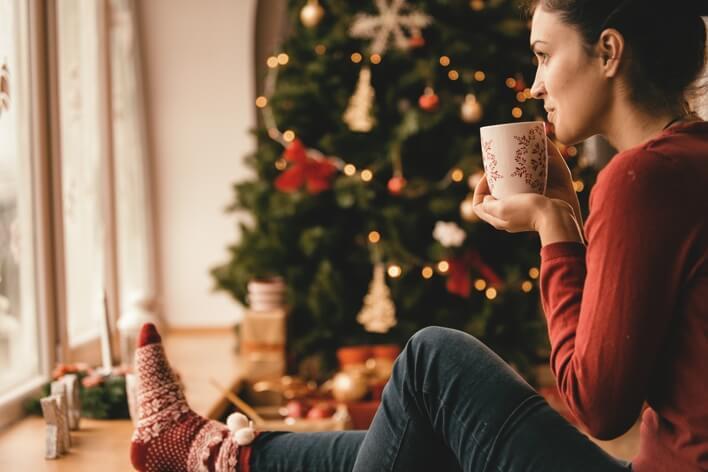 Woman in a red sweater and patterned socks, seated by a window and holding a mug, gazes outside with a decorated Christmas tree in the background, finding solace after her visit to the nearby medical center.
