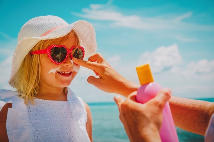 A child with heart-shaped sunglasses and a sun hat has sunscreen applied to their face at the beach, ready for fun under the sun. Remember, proper precautions can prevent medical visits or trips to urgent care for skin issues.