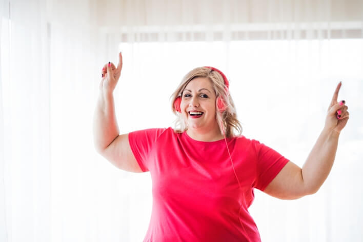 A person wearing a pink shirt and headphones smiles and points upwards with both hands in a brightly lit room, perhaps celebrating good news from their recent visit to the medical center.
