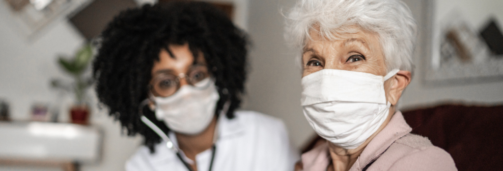 An elderly woman wearing a mask sits in the foreground at a medical center, while a masked healthcare worker attends to her in the background.