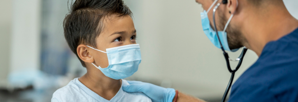 A child wearing a face mask is being examined by a healthcare professional at an urgent care clinic. The healthcare worker, also masked, uses a stethoscope to check the child's health.
