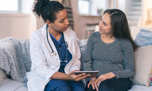 A healthcare professional in a white coat discusses medical information on a tablet with a pregnant woman in a striped shirt. They are sitting on a couch in the well-lit room of the walk-in clinic.