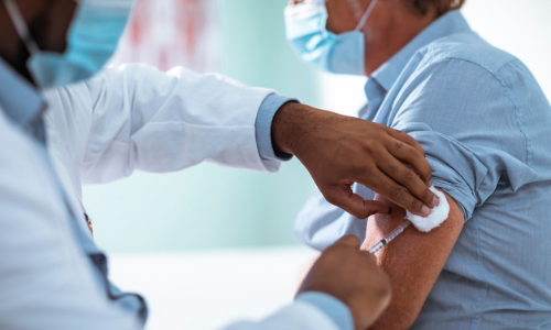 A healthcare professional at a walk-in clinic administers a vaccine to a seated adult, who is wearing a face mask and blue shirt. The professional, clad in a white coat and also wearing a face mask, provides urgent care with precision and compassion.