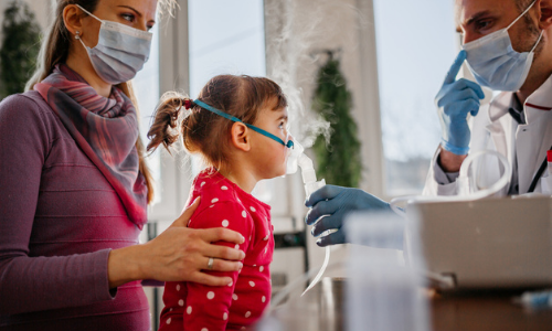 A child wearing a red polka-dot shirt uses a nebulizer mask, assisted by a doctor in an urgent care clinic and accompanied by an adult, both wearing masks.