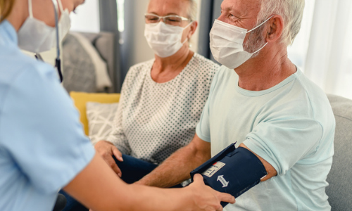 A healthcare worker at a medical center measures the blood pressure of an elderly man wearing a face mask, while an elderly woman, also masked, sits beside him.
