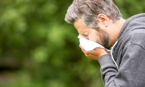 A man with gray hair and a beard is sneezing or coughing into a tissue while standing outdoors, wearing a dark hoodie, possibly on his way to a medical center.