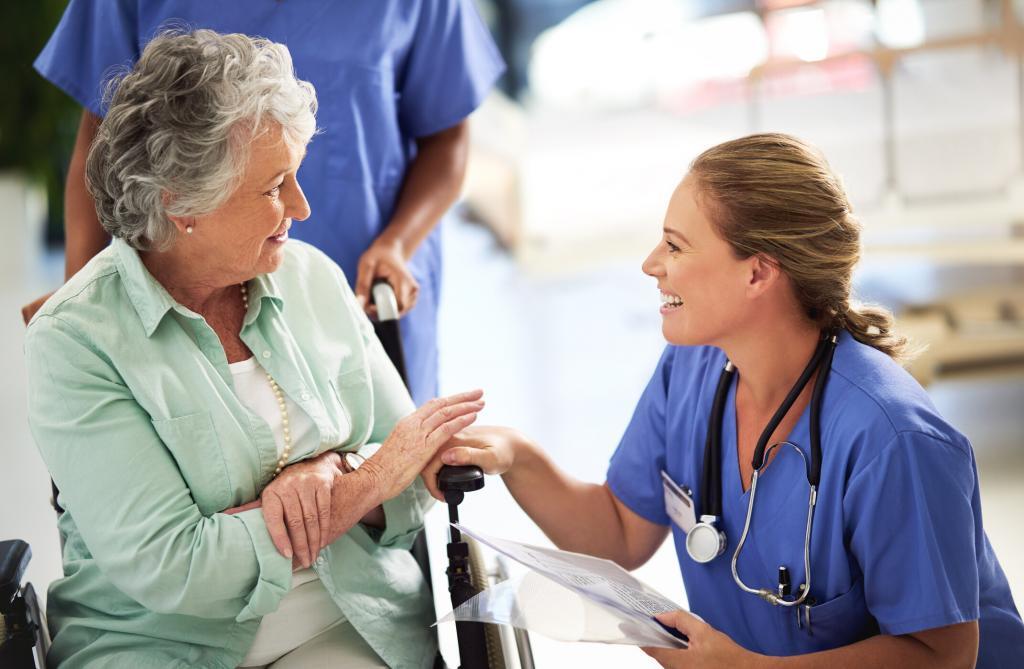 Young nurse in blue scrubs smiling and holding a clipboard, interacting with a seated elderly woman in light-colored clothing, with another medical professional in the background at an urgent care center.